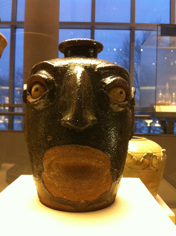 Face Jug. Maker unknown. Western South Carolina. Ca. 1860-70. Alkaline-glazed stoneware. This jug and others similar to it were made by African American slaves and freedmen – one of the former signed his pieces “Dave the Slave” – working in potteries in and around Edgefield, along the Horse Creek Valley, rich in clay deposits. It is said the style and material of these vessels relate to ritualistic objects of the Kongo and Angola. Most likely, the face lost its teeth and lips during the firing process.